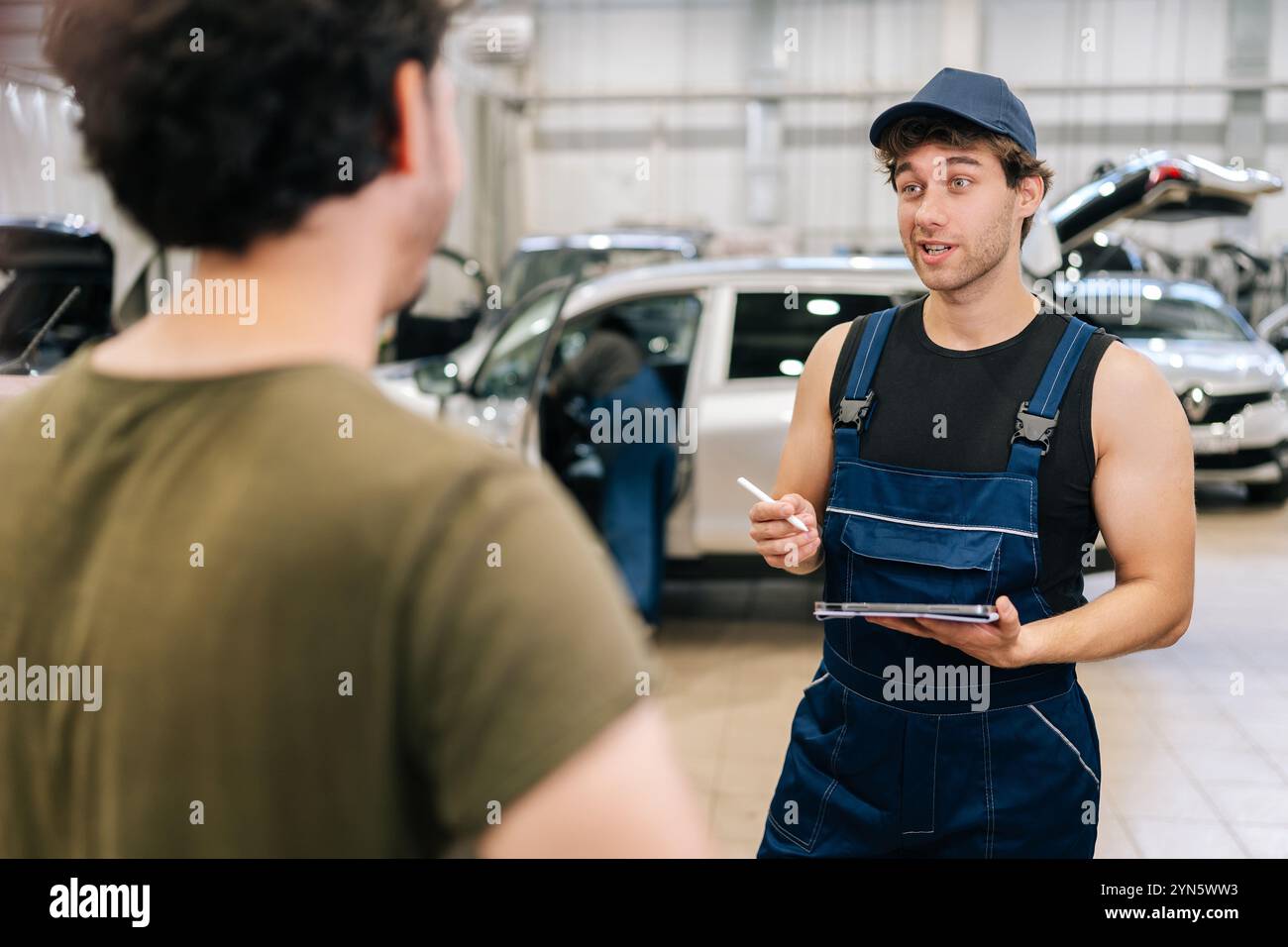 Travailleur de garage masculin en uniforme et client homme méconnaissable parlant pour discuter des détails des réparations de voiture. Mécanicien automatique tenant la tablette et écrivant Banque D'Images