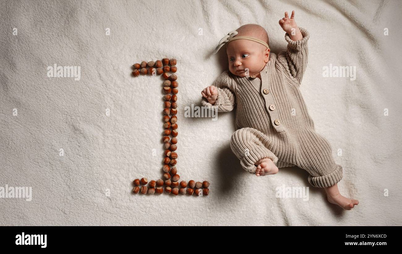 Bébé européen actif en pleine hauteur, âgé d'un mois, sur une couverture dans une salopette tricotée avec un arc sur la tête, le numéro est celui des noix naturelles, pastel Banque D'Images