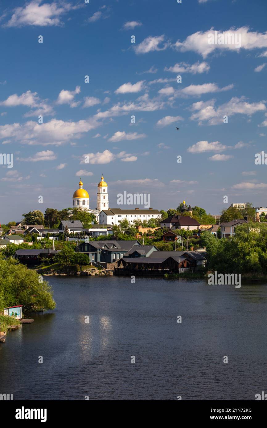 Une scène sereine au bord de la rivière avec une église au dôme doré, des maisons traditionnelles et une végétation luxuriante sous un ciel bleu vif avec des nuages éparpillés, se reflètent Banque D'Images
