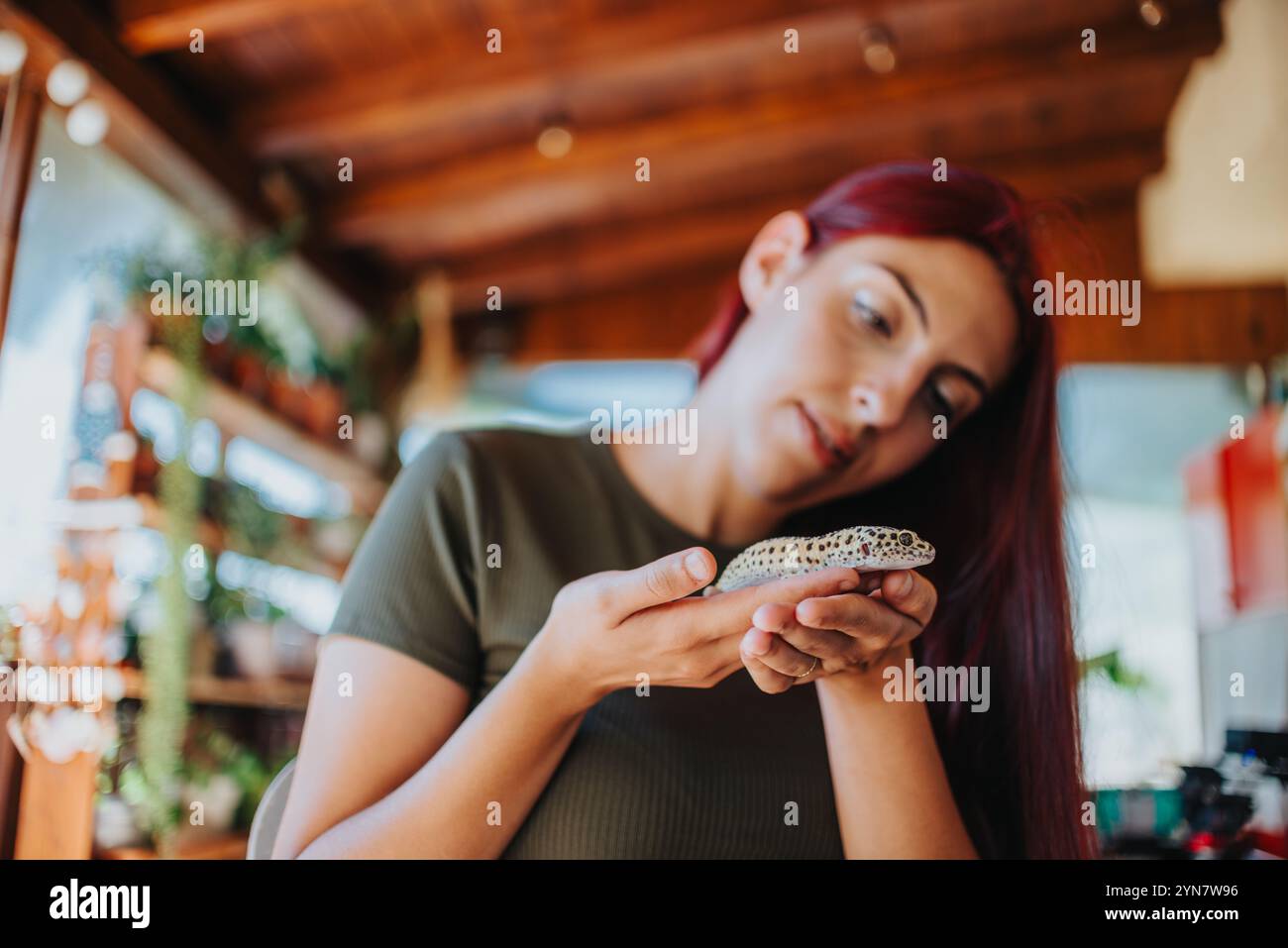 Jeune femme tenant doucement un lézard dans ses mains tout en étant assise dans un espace intérieur confortable rempli de verdure et de décor en bois Banque D'Images