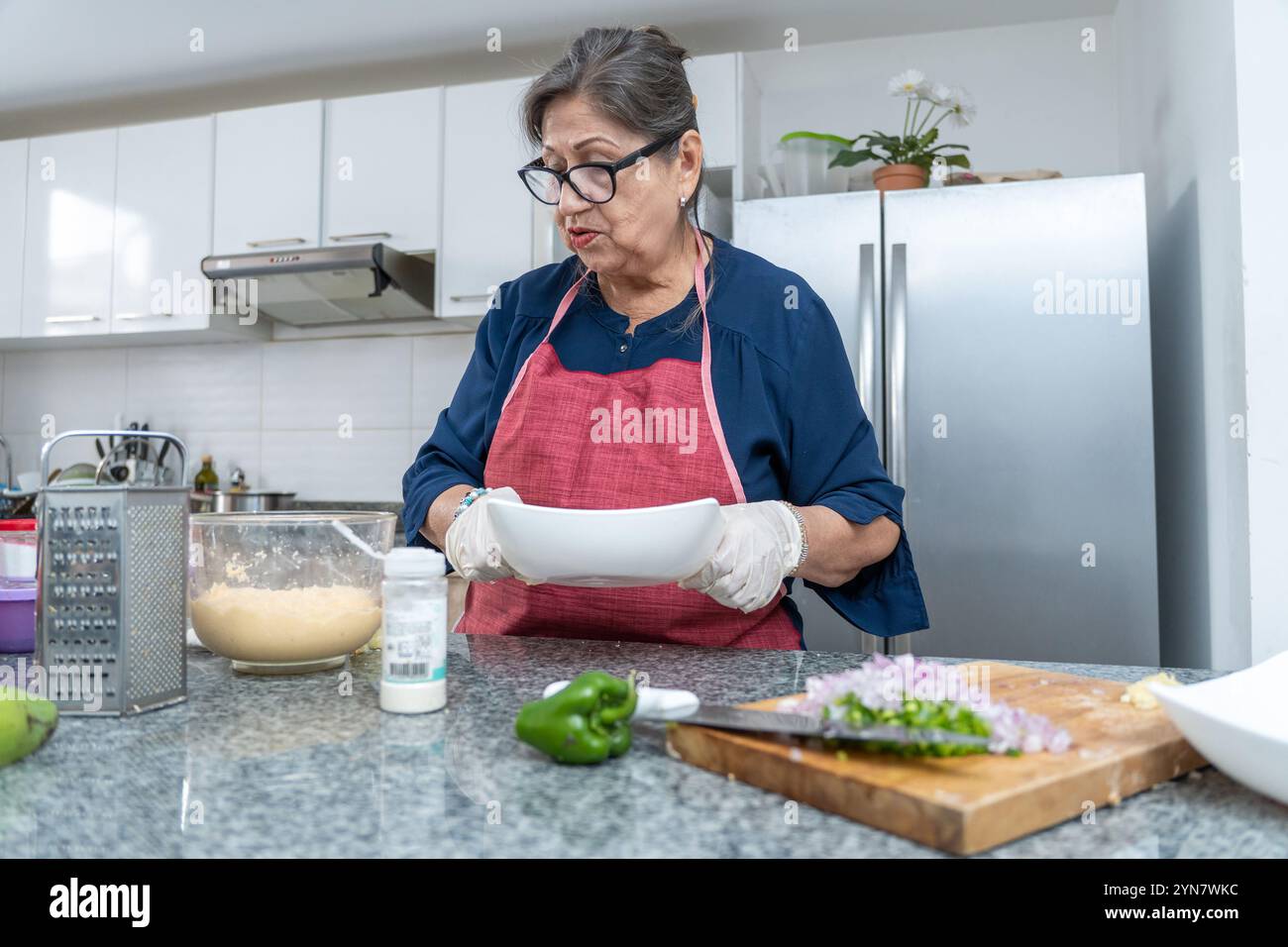 Une femme dans un tablier bleu prépare de la nourriture dans une cuisine. Elle porte des gants et tient une assiette blanche Banque D'Images