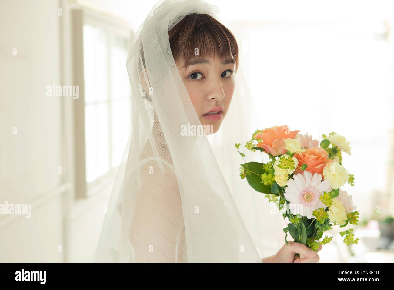 Femme dans sa vingtaine en robe de mariée tenant un bouquet par la fenêtre Banque D'Images
