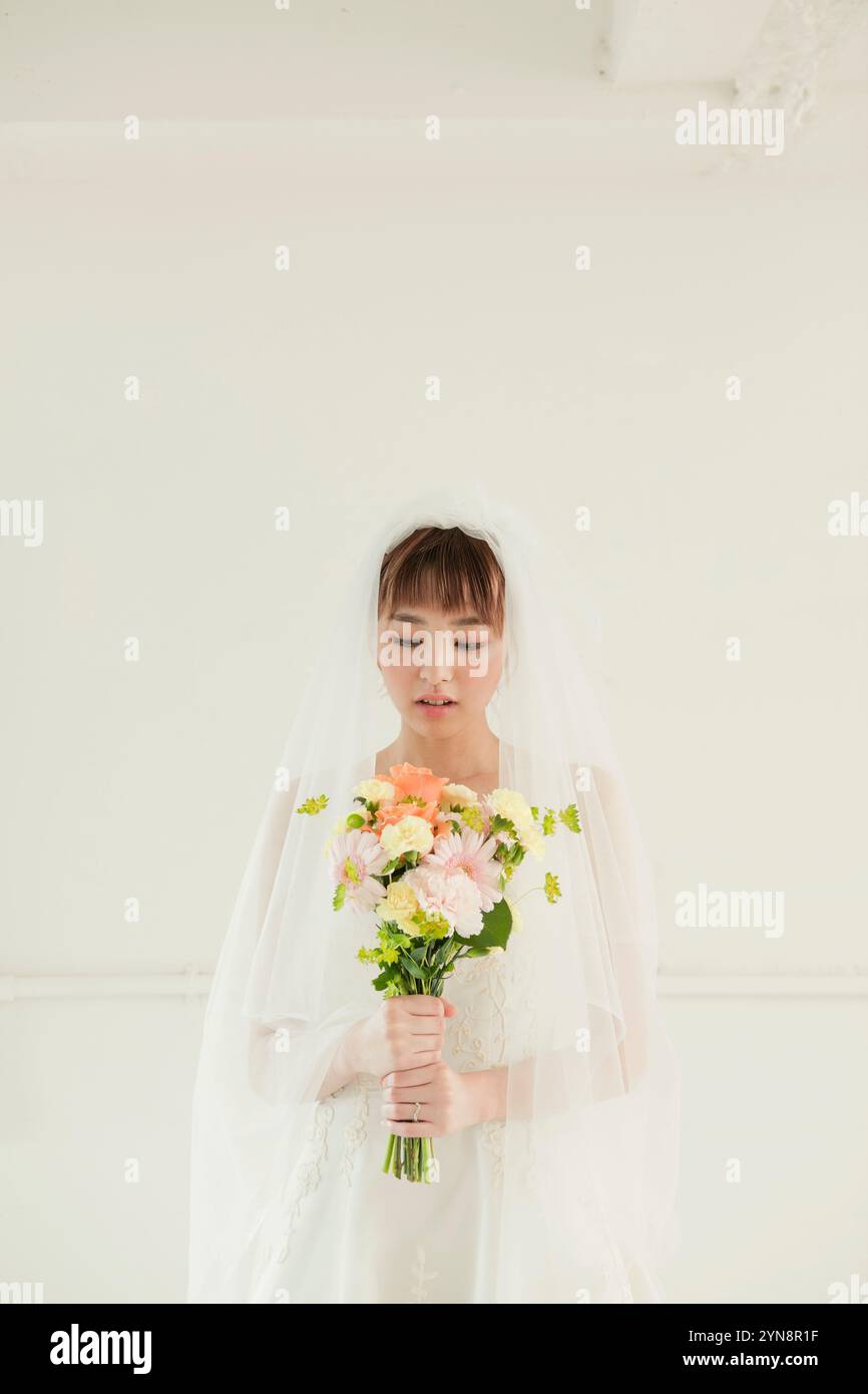 Femme dans sa vingtaine en robe de mariée tenant un bouquet par la fenêtre Banque D'Images
