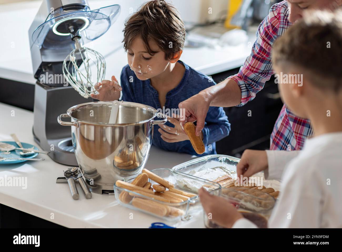 Deux enfants participent joyeusement à une activité de cuisson, mélangeant les ingrédients avec un batteur électrique dans une cuisine lumineuse pendant que l'on goûte la pâte. Banque D'Images