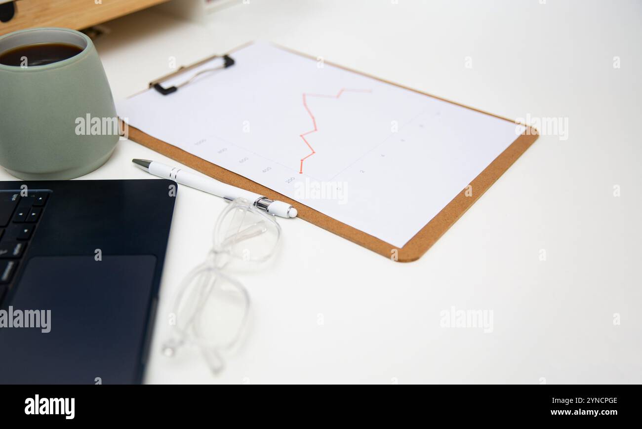 Table de travail de bureau avec rapport de graphiques sur presse-papiers en bois, café, fournitures et ordinateur portable. Banque D'Images