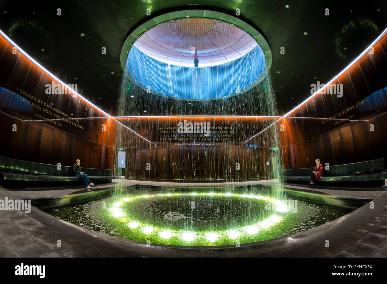 Musée national d'histoire et de culture afro-américaines Cour contemplative Washington DC | WASHINGTON D.C., États-Unis — Une vue fisheye capture la cour contemplative du musée national d'histoire et de culture afro-américaines, où une fontaine cylindrique tombe d'un oculus éclairé dans une piscine immobile. Les murs en verre caramel-bronze de Bendheim, gravés de citations de Nelson Mandela et Martin Luther King Jr., entourent l’espace Concourse Level 0 conçu par Philip Freelon et David Adjaye. Le musée a ouvert ses portes le 24 septembre 2016 en tant que 19e musée du Smithsonian. Banque D'Images