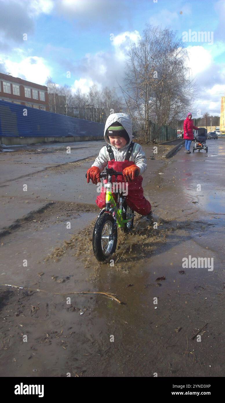 Un tout-petit vêtu de vêtements chauds d'hiver roule heureux sur un vélo d'équilibre à travers une flaque, éclaboussant de joie. Sa mère regarde de loin avec un sourire, tenant Banque D'Images