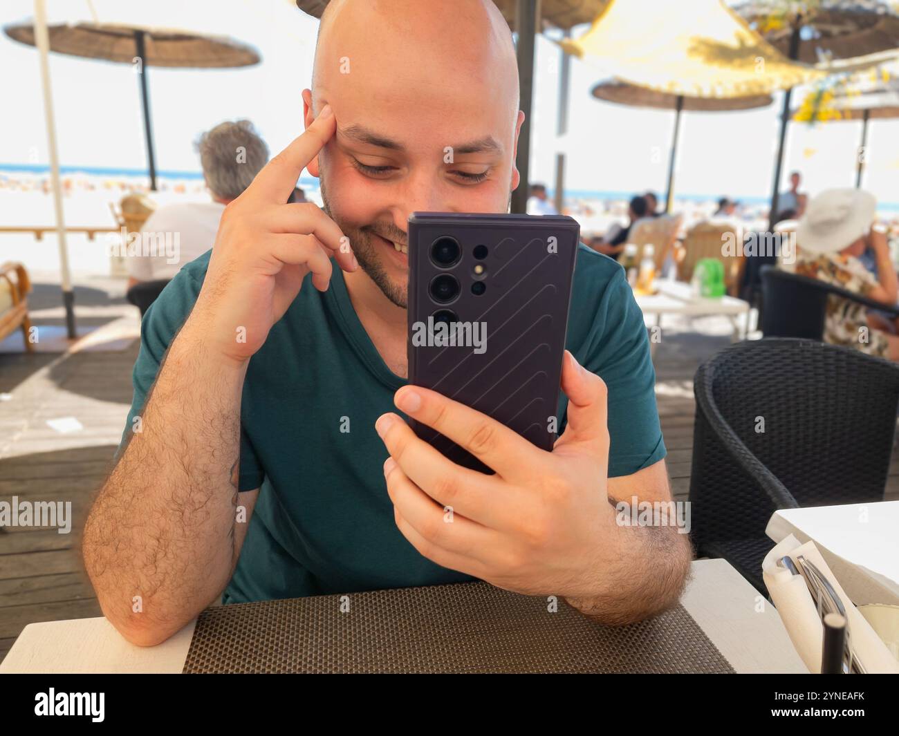 Jeune homme souriant utilisant son téléphone portable dans un restaurant de plage par une journée ensoleillée, regardant le téléphone portable rire - Image de stock capturée avec un smartphone