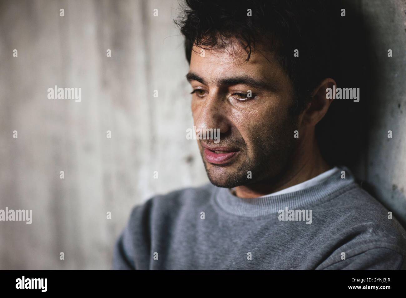 Un homme aux cheveux touffus s'assied tranquillement contre un mur texturé, perdu dans ses pensées. Son expression révèle la contemplation, créant un sentiment d’introspection Banque D'Images