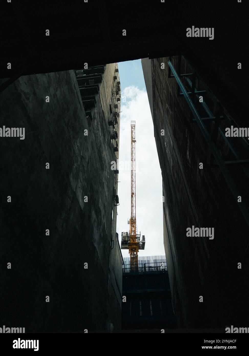 Grue de construction encadrée par de hauts murs urbains dans une ruelle étroite, capturant le contraste du progrès industriel par rapport à l'architecture Stark Banque D'Images
