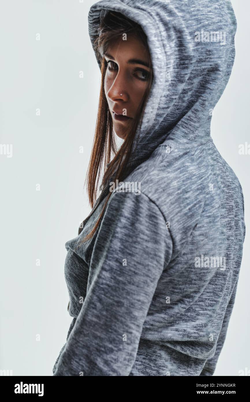 Jeune femme élégante dans un sweat à capuche gris regarde en arrière avec confiance et mystère, exsudant la beauté urbaine moderne Banque D'Images