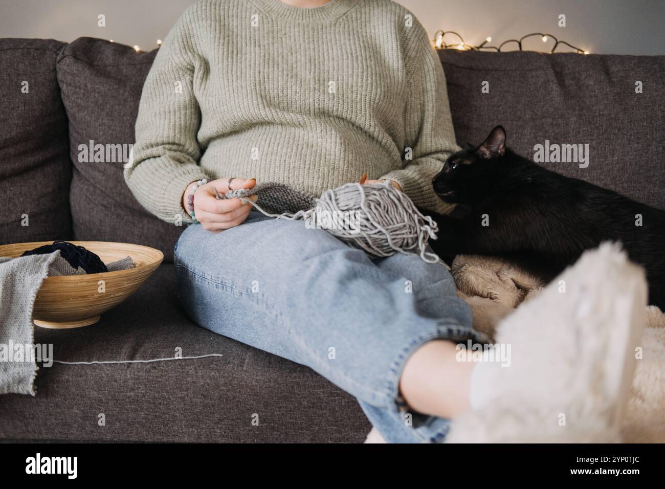 Femme tricotant avec chat noir sur canapé, embrassant temps d'arrêt confortable. Relaxation, compagnie, détox numérique et concept de passe-temps attentif. Banque D'Images