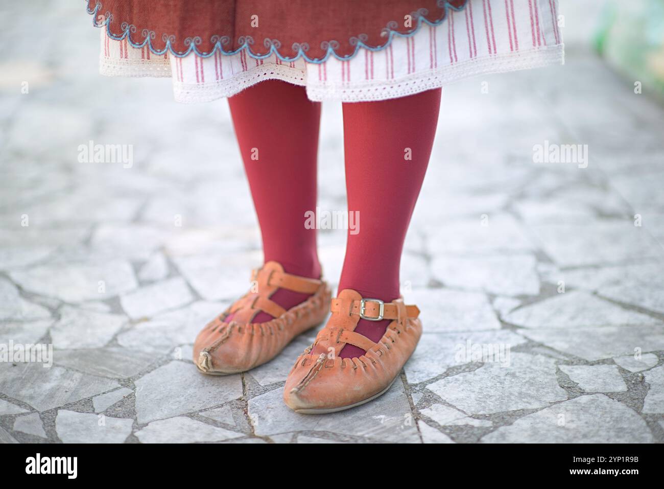 Costume traditionnel authentique de la région des Shopes avec bas rouge vif, ourlet de jupe brodé et chaussures en cuir distinctives sur pavé. Banque D'Images