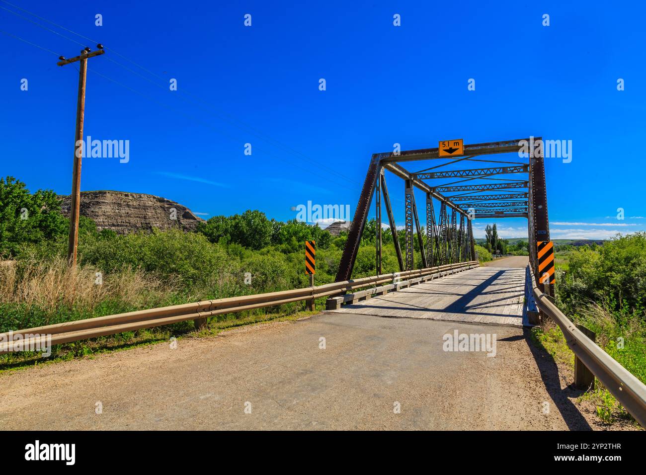 Un pont enjambe une route au-dessus d'un champ herbeux. Le pont est vieux et a un panneau jaune et noir dessus. Le ciel est bleu et clair Banque D'Images