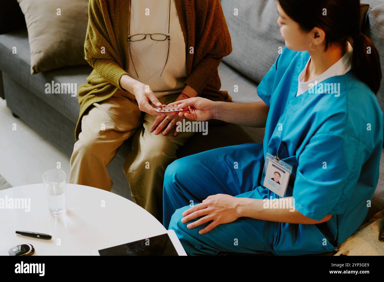 Infirmière portant des gommages bleus aidant la femme âgée avec des médicaments tout en étant assise sur le canapé dans le cadre confortable du salon avec table basse au premier plan Banque D'Images