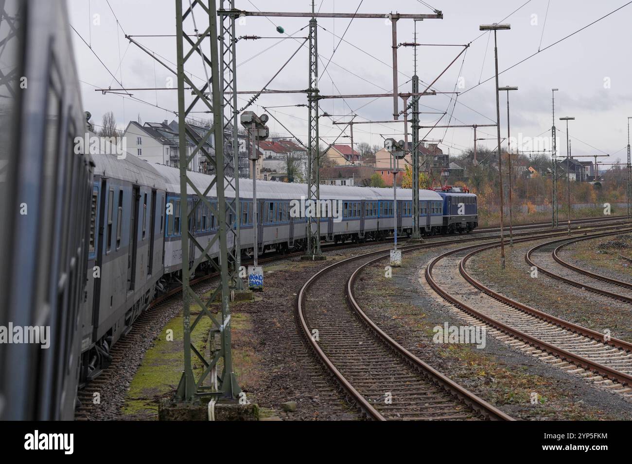 Historischer Zug mit blauen waggons auf kurviger Gleisanlage, umgeben von Oberleitungen und Signalmasten, weitläufiges Gleisnetz, herstliche Vegetation am Rand, Städtische Bebauung im Hintergrund, typisches Bild des regionalen Bahnverkehrs, Verbindung von nostalgischem Bahnverkehr und moderner Infrastruktur Schienenführung Banque D'Images