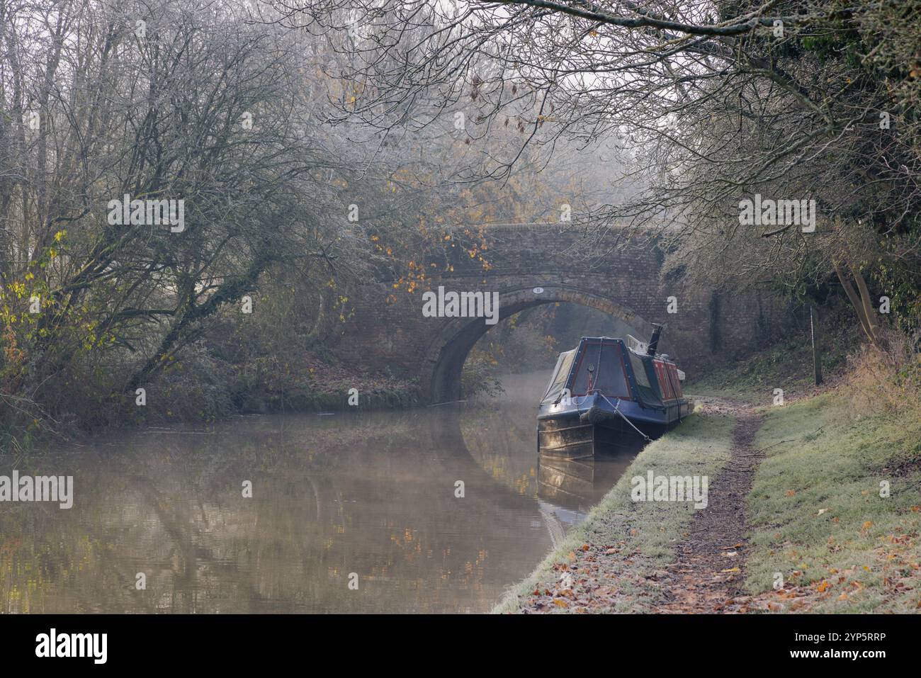 Crick, Northamptonshire, Royaume-Uni, 2024 : un matin brumeux, en fin d'automne, un bateau étroit est amarré sur le canal Grand Union par un vieux pont à bosse. Banque D'Images