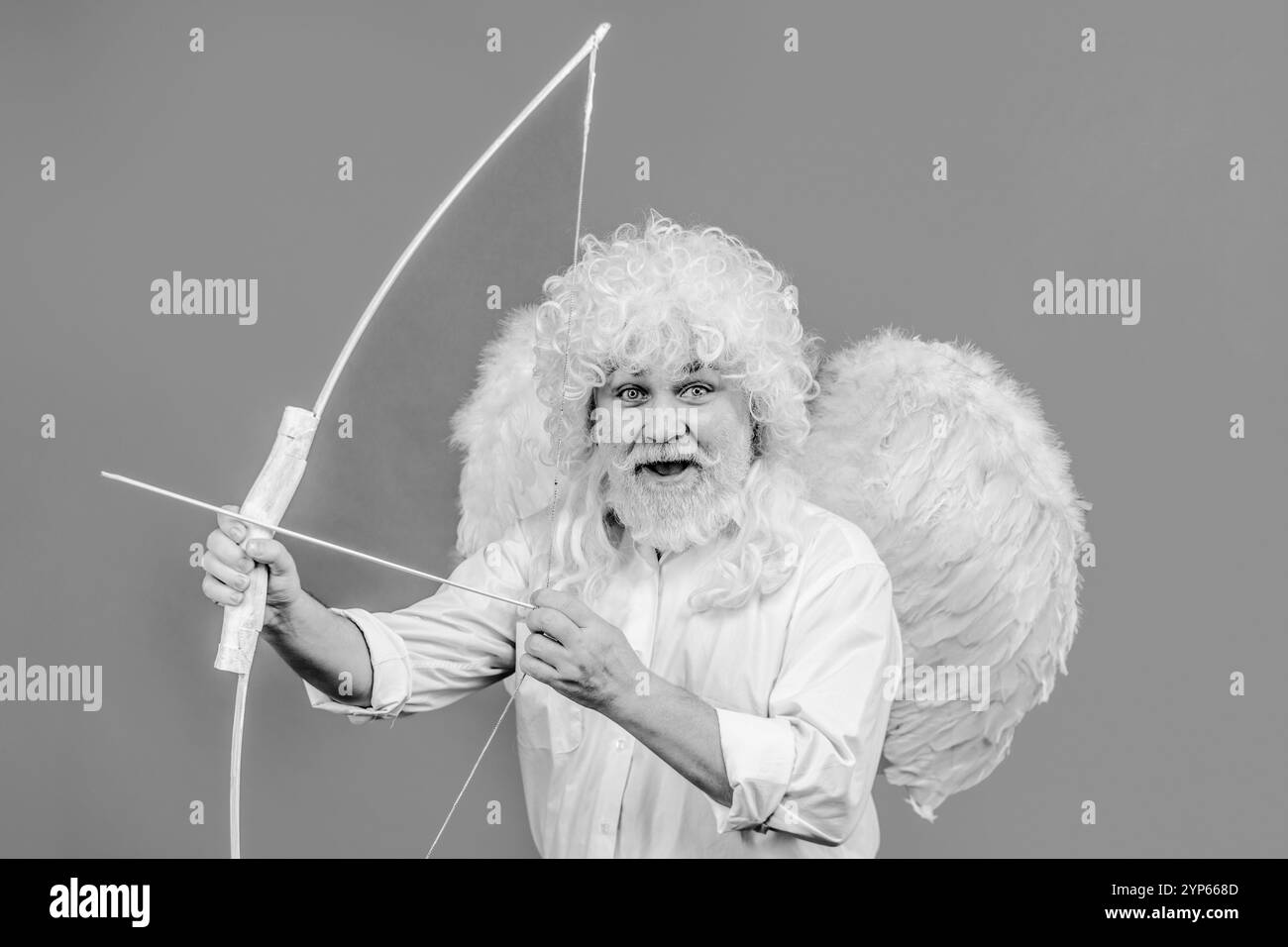 Célébration de la Saint-Valentin. Heureux homme barbu avec des ailes d'ange tirant flèche d'amour. Bel homme en costume d'ange et perruque bouclée avec arc et flèches Banque D'Images