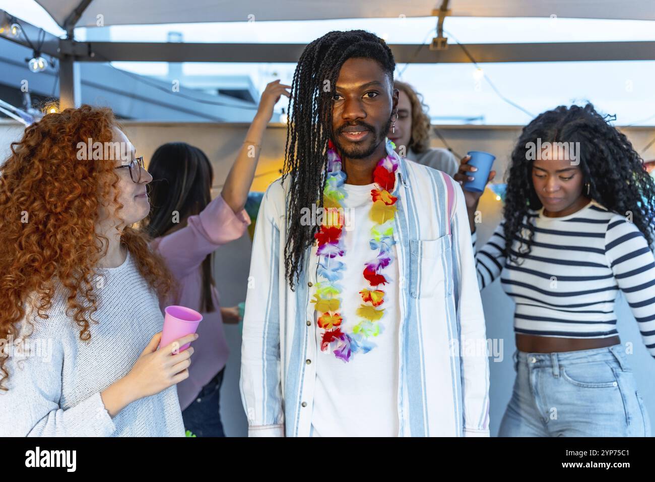 Homme avec coiffure afro et collier hawaïen debout avec des amis multiraciaux à la fête sur le toit Banque D'Images