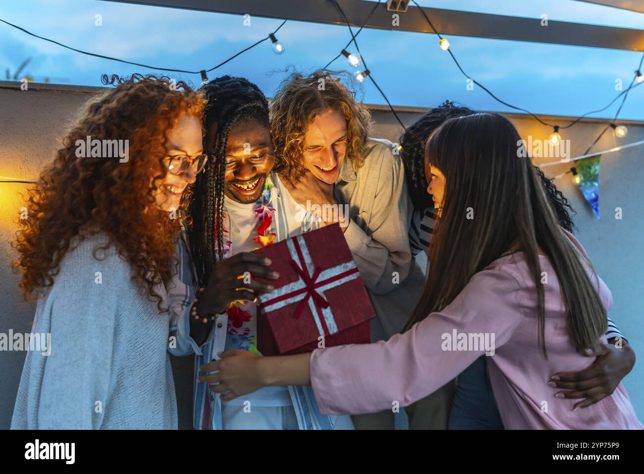 Homme africain ouvrant cadeau d'anniversaire embrassé par ses amis multiraciaux lors de la fête sur le toit Banque D'Images