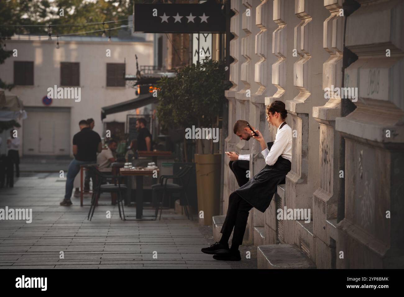 BELGRADE, SERBIE - 12 JUIN 2024 : deux serveurs, un homme et une femme, assis à l'extérieur sur une pause et regardant leurs smartphones illustrant le tec moderne Banque D'Images