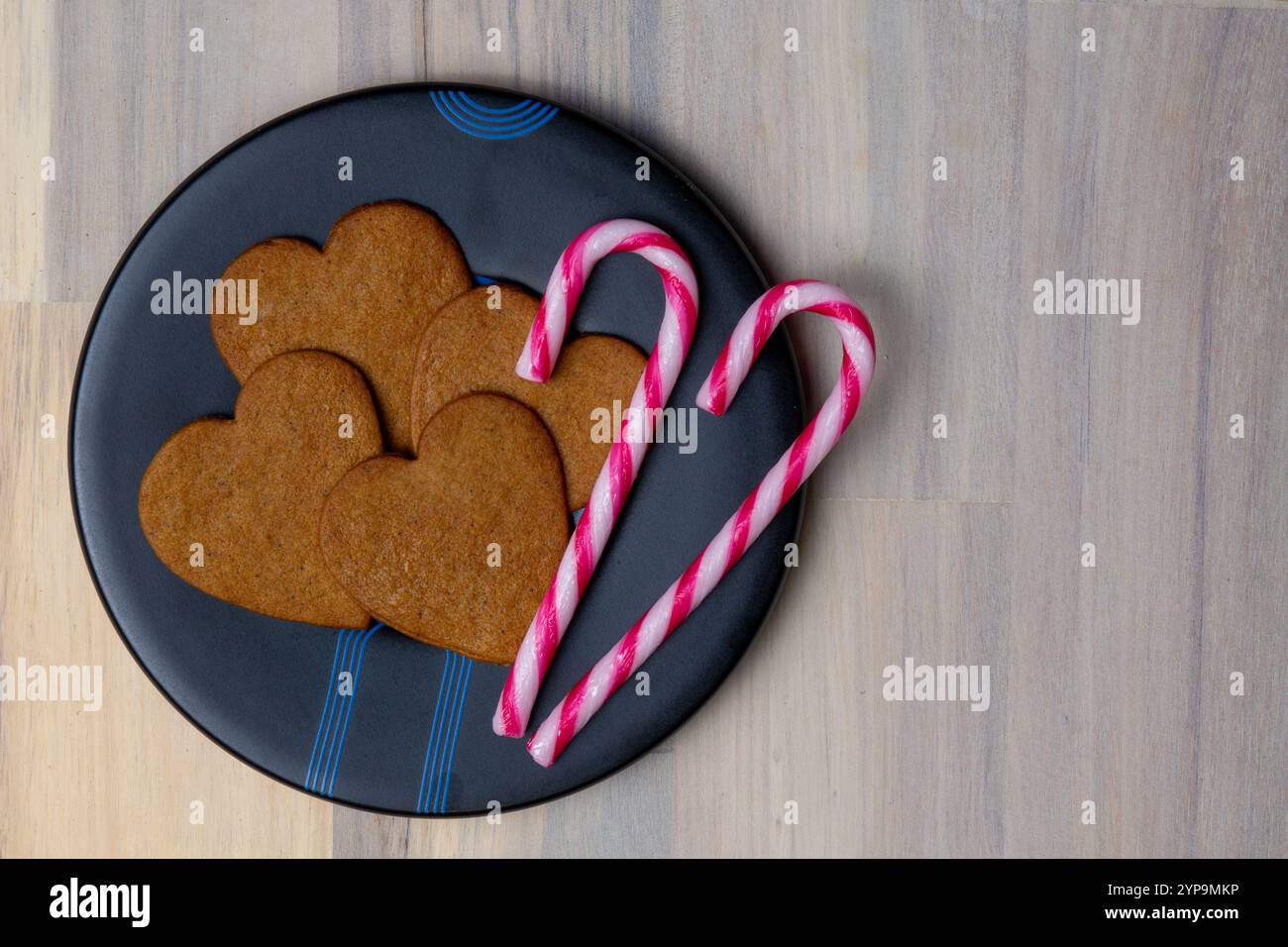 Des biscuits en forme de cœur en pain d'épice et des cannes de bonbon rayées roses sont disposés sur une assiette noire, créant un affichage festif et invitant. Banque D'Images