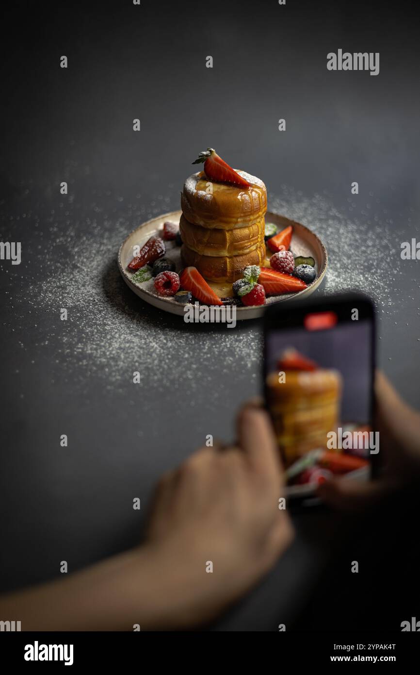 Personne photographiant une assiette de crêpes japonaises moelleuses avec des baies fraîches, du sucre en poudre et du sirop à l'aide d'un smartphone Banque D'Images