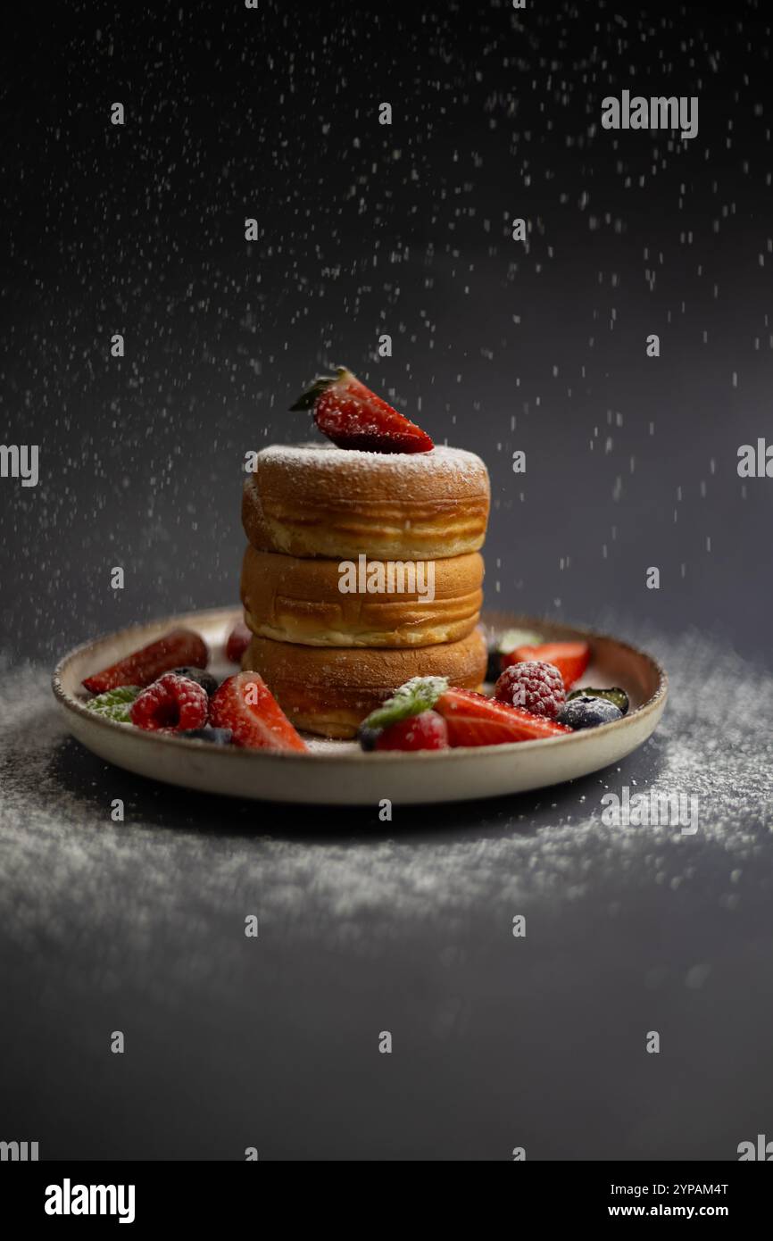 Pile de crêpes japonaises moelleuses avec des fraises fraîches, des framboises, des bleuets et de la menthe sur une assiette, surmontée de sucre en poudre tombant comme de la neige Banque D'Images