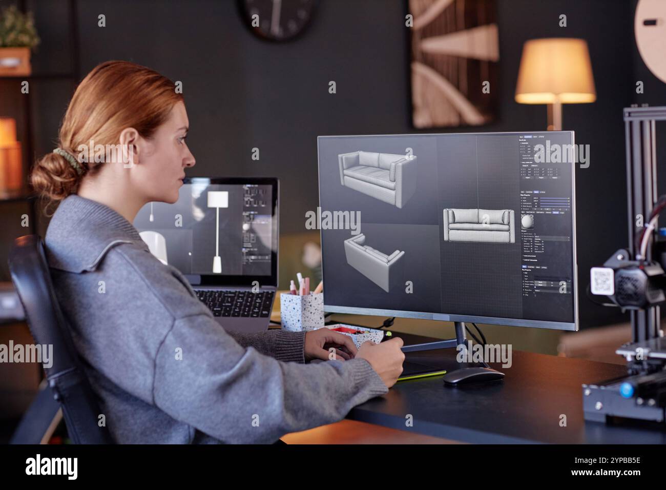 Femme assise au bureau, se concentrant sur un logiciel de modélisation 3D affiché sur son écran d'ordinateur. L'environnement de bureau comprend divers gadgets technologiques et des éléments de décoration modernes Banque D'Images