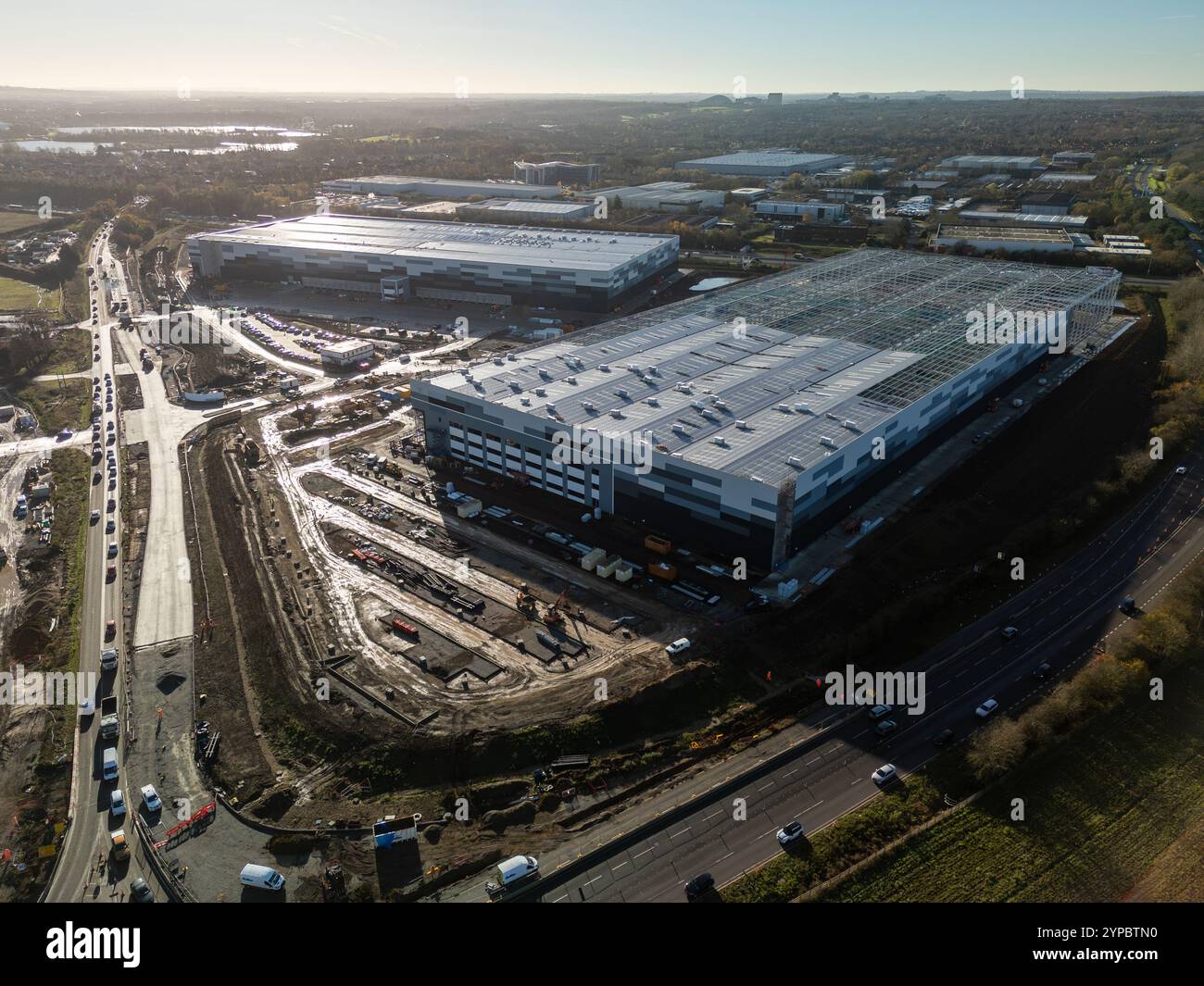 Vue aérienne d'un grand entrepôt de distribution en construction à côté d'une autoroute très fréquentée, montrant l'échelle de la logistique moderne Banque D'Images