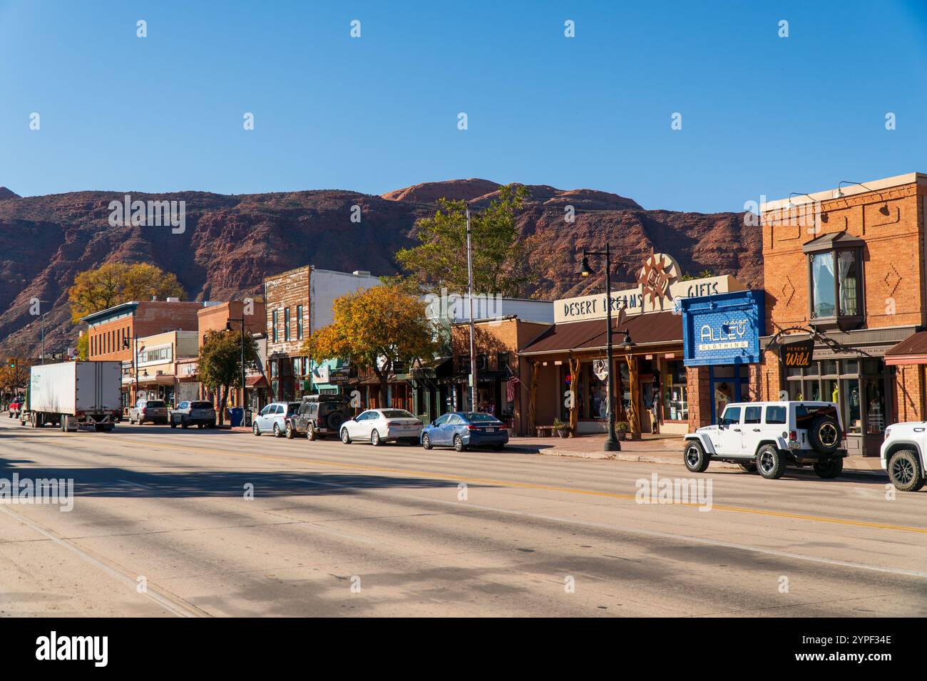 Rue principale dans le centre-ville de Moab, Utah, montrant des devantures historiques, des boutiques de souvenirs, des véhicules garés et des falaises de roche rouge sous un ciel bleu clair Banque D'Images