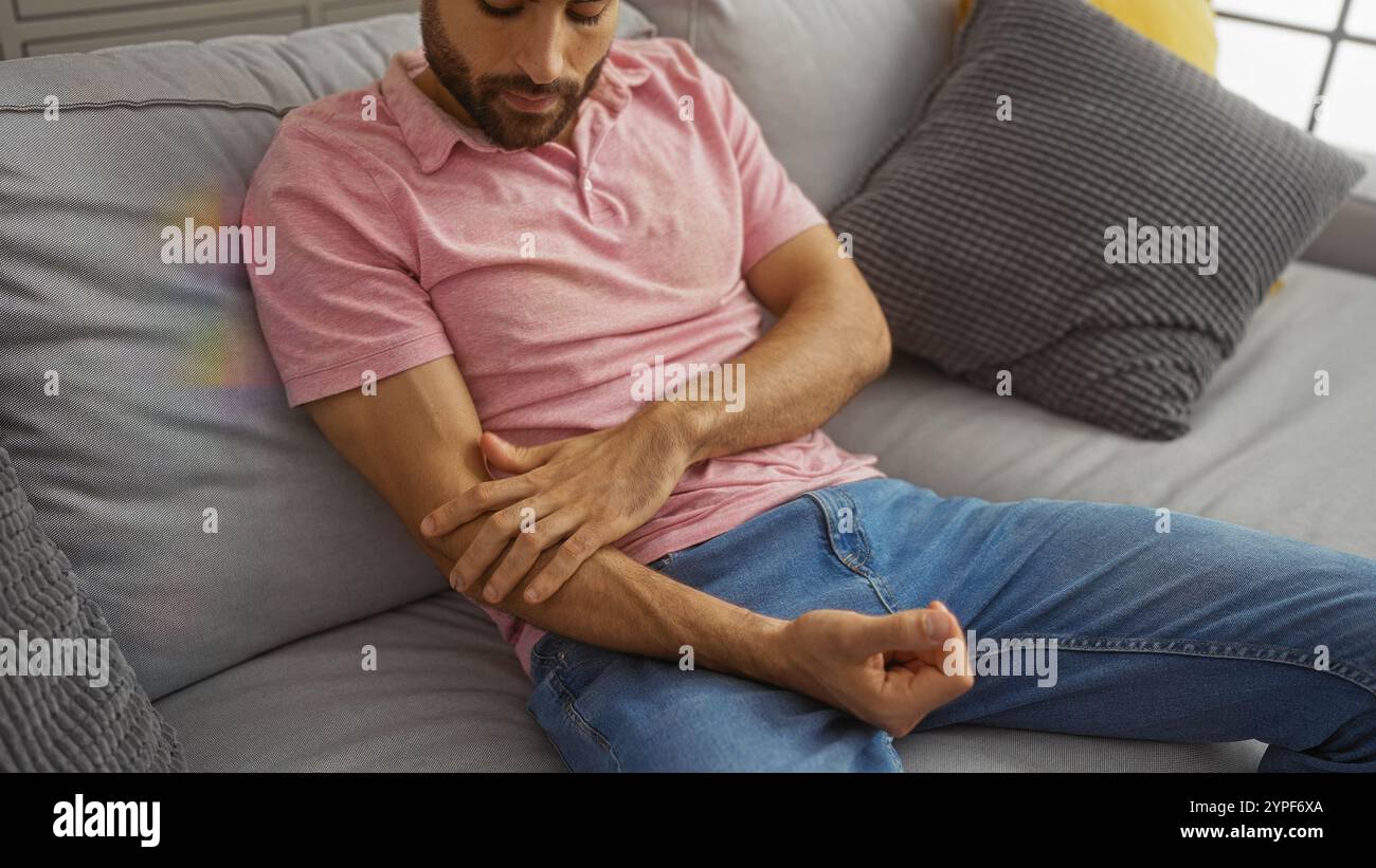 Jeune homme hispanique est assis sur un canapé confortable à l'intérieur d'un appartement moderne tout en touchant son coude doucement Banque D'Images