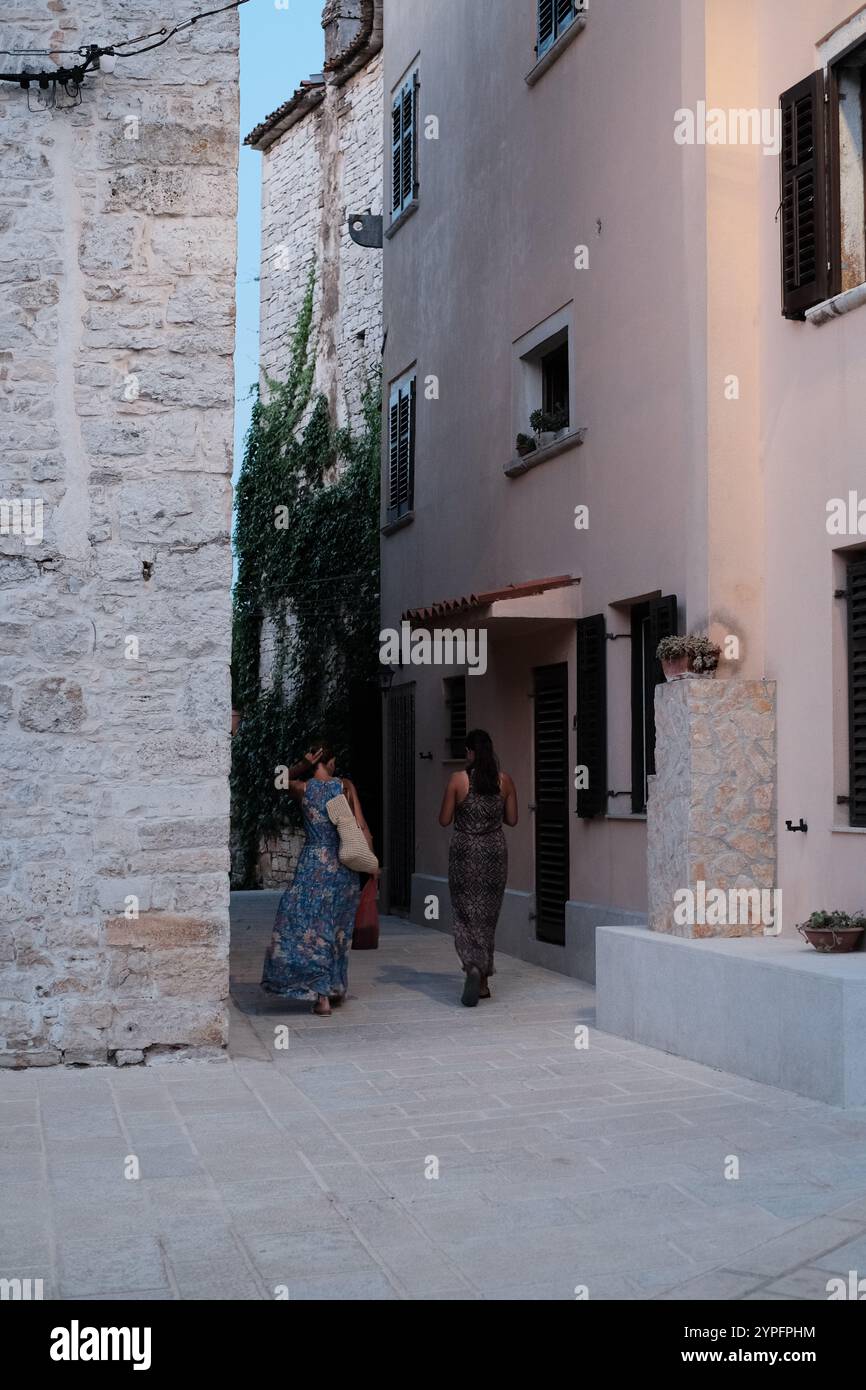 Deux jeunes femmes en robes colorées marchant dans la vieille ville. Maisons en pierre et ruelle étroite à Bale, Istrie. Banque D'Images