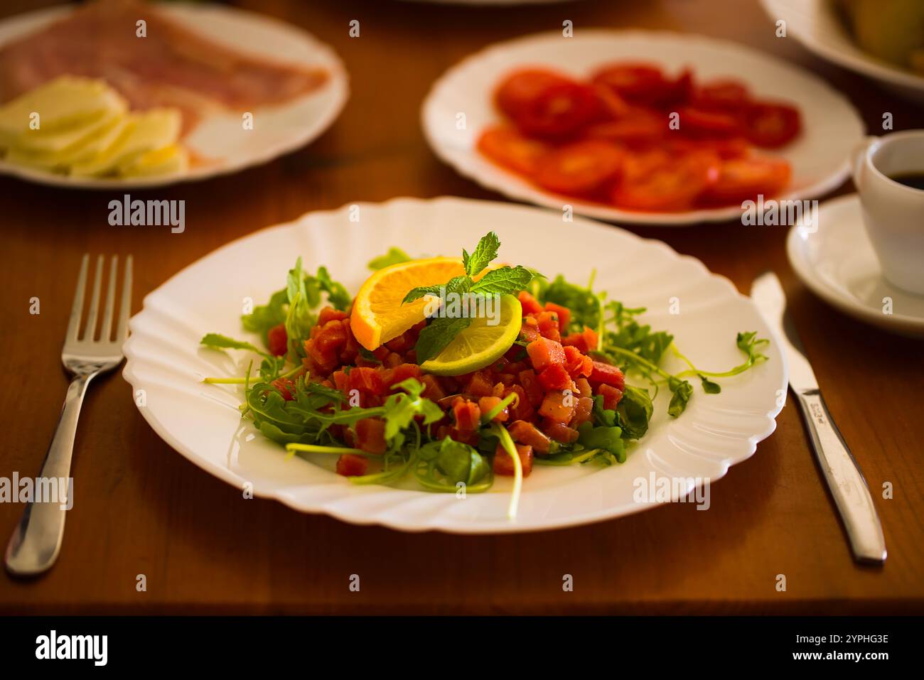 Tartare de thon frais sur une assiette blanche, décoré de roquette, menthe et tranches d'agrumes, se trouve au sommet d'une table en bois, entouré d'autres assiettes de petit déjeuner i. Banque D'Images