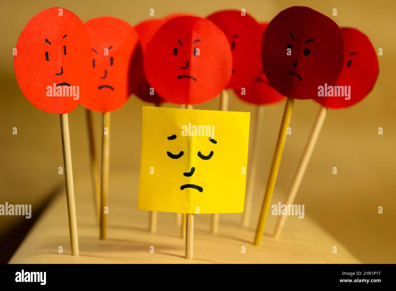Image horizontale de papier jaune avec un visage triste entouré de visages en colère, concept de rejet social et de discrimination Banque D'Images