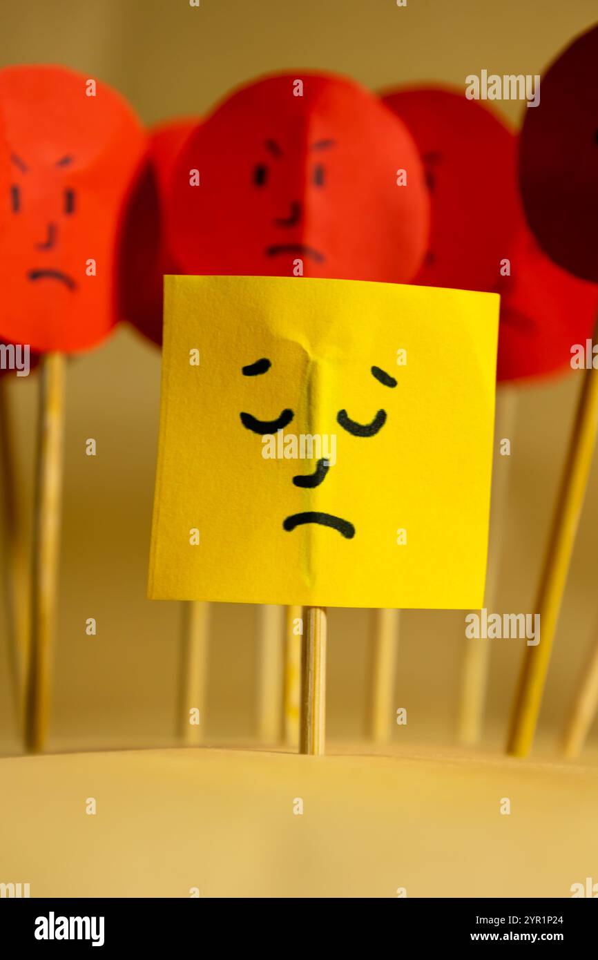 Image verticale de papier jaune avec un visage triste entouré de visages en colère, concept de rejet social et de discrimination Banque D'Images