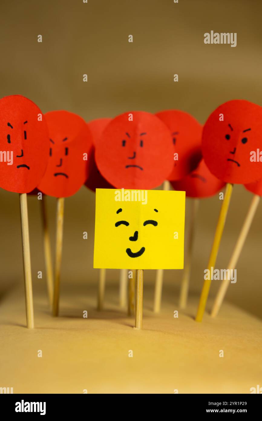 Image verticale de papier jaune avec un visage souriant entouré de visages en colère, concept de rejet social et de discrimination Banque D'Images