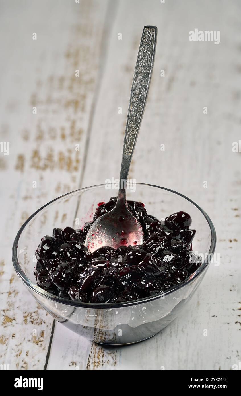 Confiture noire sauvage amère de cerises douces dans un petit bol en verre avec une cuillère à café, sur un fond en bois rustique Banque D'Images