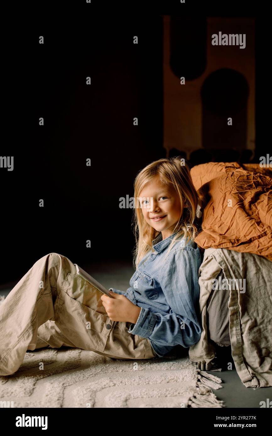 Portrait de garçon souriant assis avec livre près du lit sur le sol dans la chambre à la maison Banque D'Images