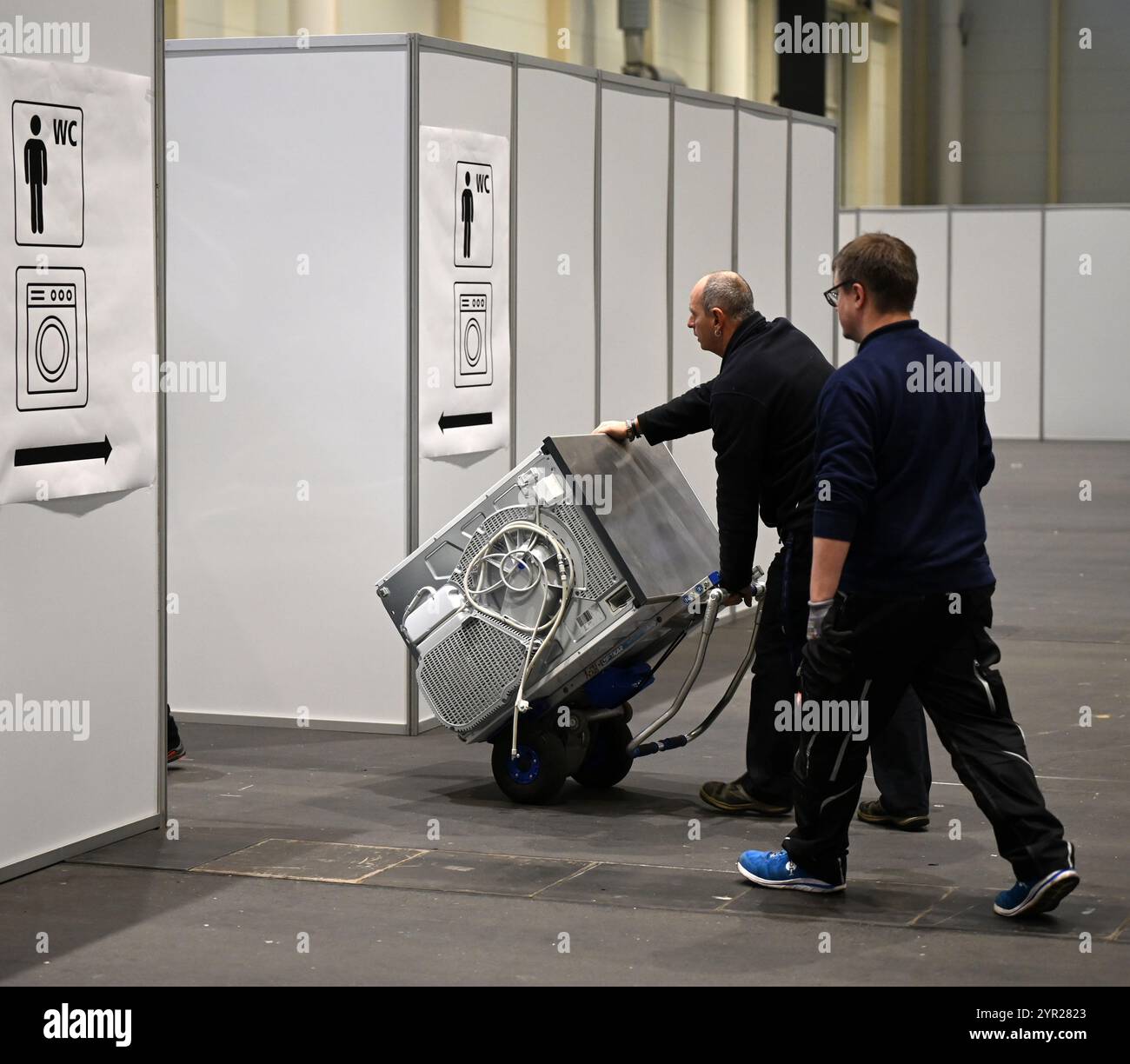 02 décembre 2024, Hambourg : deux ouvriers poussent une machine à laver dans les installations de lavage lors d'un rendez-vous avec les médias dans le logement des réfugiés dans le hall d'exposition B5 sur le terrain d'exposition. À partir du 3 décembre, le Hall B5 deviendra à nouveau un hébergement d’urgence pour les réfugiés. L'occupation est prévue pour jusqu'à 300 personnes, avec une capacité d'urgence totale de 476 places dans les compartiments. Le logement sera géré par l’entreprise sociale municipale Fördern & Wohnen. Photo : Niklas Graeber/dpa Banque D'Images