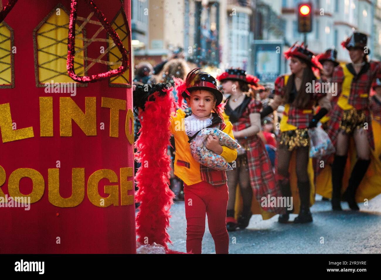 L'image montre un défilé ou un spectacle de rue en cours. Au premier plan, un enfant porte un costume de pompier, debout à côté d'un panneau rouge Banque D'Images