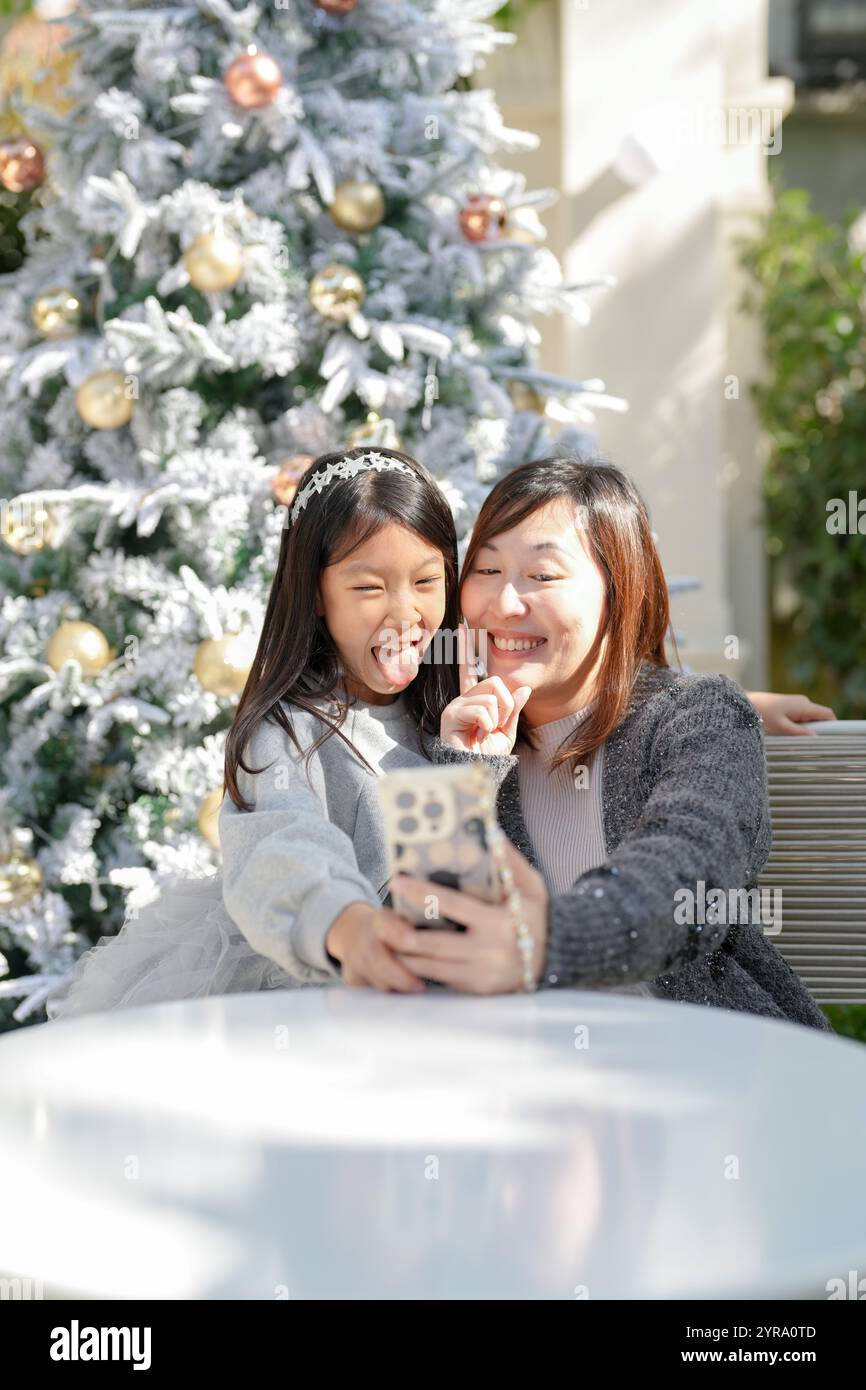 Femme singapourienne dans ses 40 ans et sa fille de 9 ans prennent selfie avec smartphone avec arbre de Noël en arrière-plan dans un charmant café décoré pour CH Banque D'Images