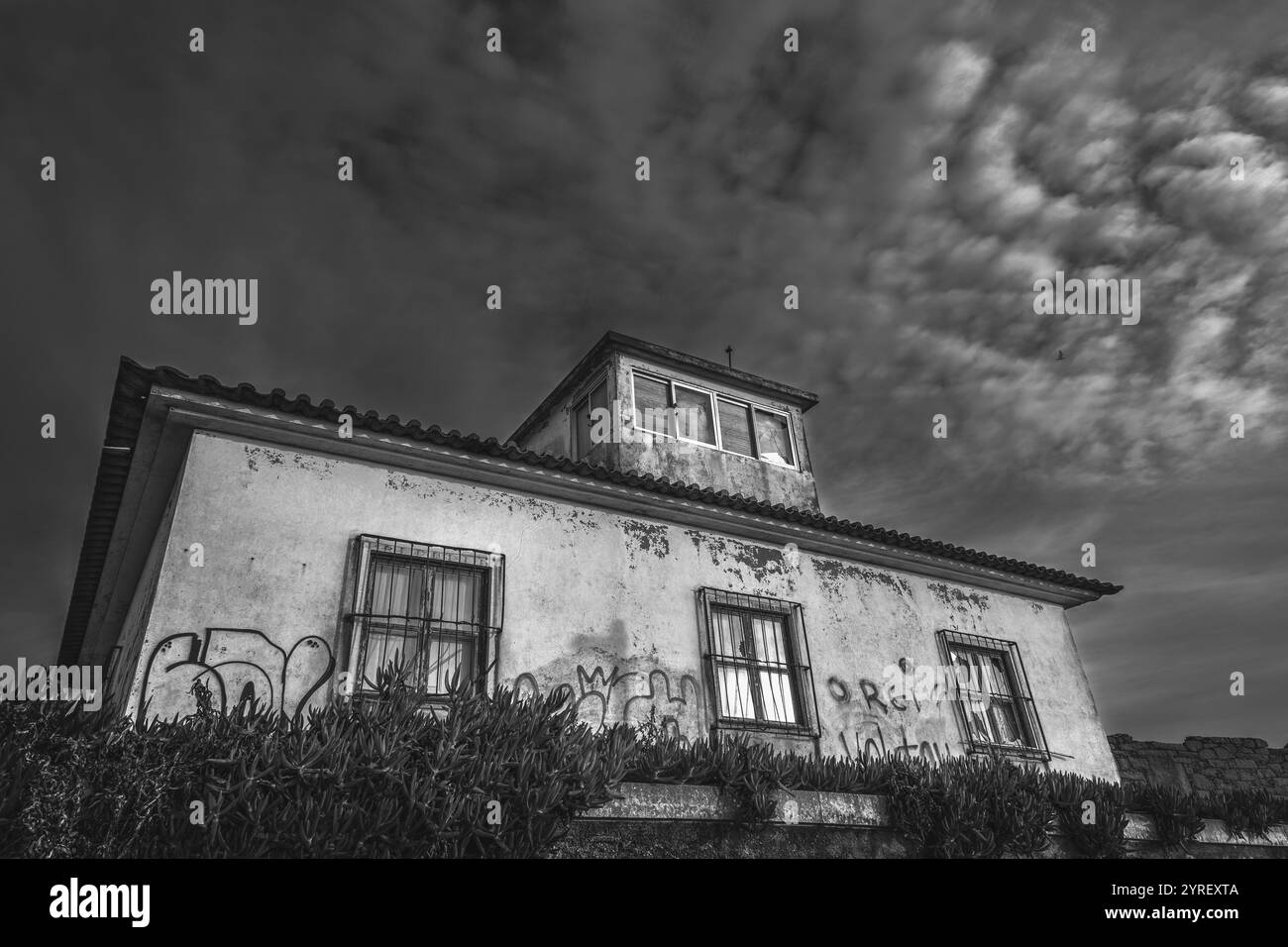 Façade en ruine d'une maison abandonnée ornée de graffitis et de fenêtres barrées, dressée contre un ciel spectaculaire et nuageux en monochrome Banque D'Images