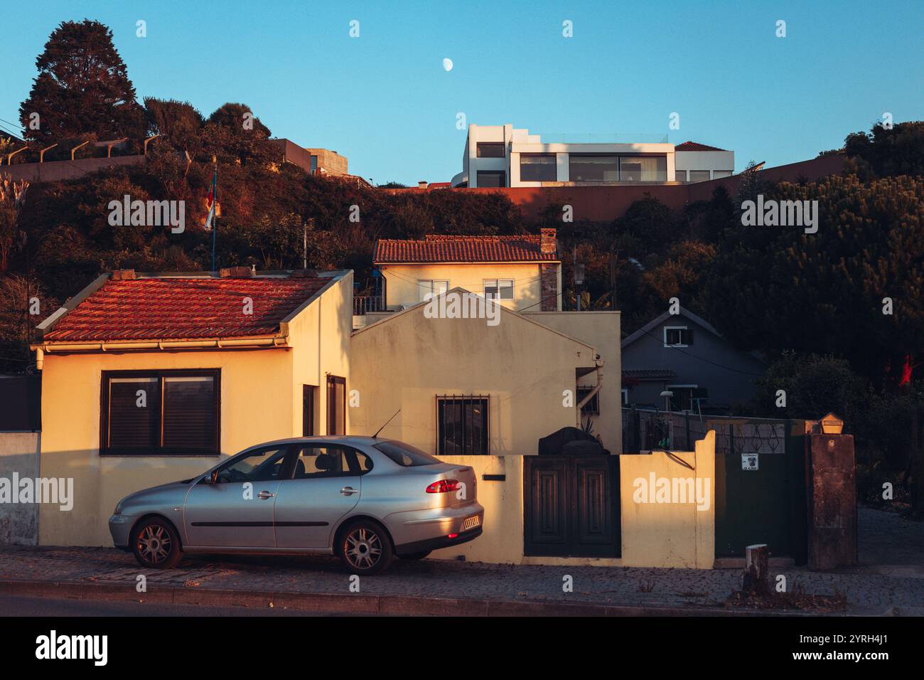 Voiture argentée garée dans la rue devant les maisons dans la lumière chaude du coucher du soleil avec la lune visible dans le ciel clair au-dessus de vila nova de gaia Banque D'Images