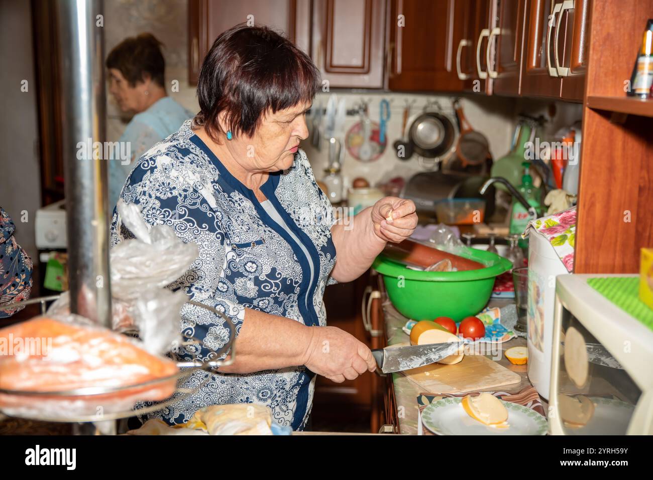 Une femme de 70 ans tranche de la saucisse sur une planche à découper en bois dans la cuisine, en essayant un morceau. Son amie aide à préparer une table festive. Le kit vintage Banque D'Images
