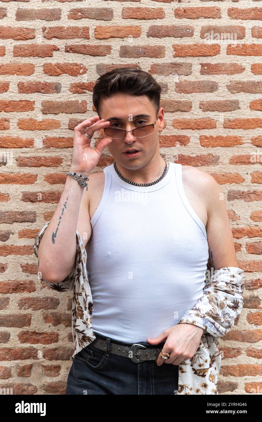 Un jeune homme à la mode dans un haut blanc sans manches et chemise à motifs ajuste ses lunettes de soleil, debout avec confiance contre un dos de mur de briques texturées Banque D'Images