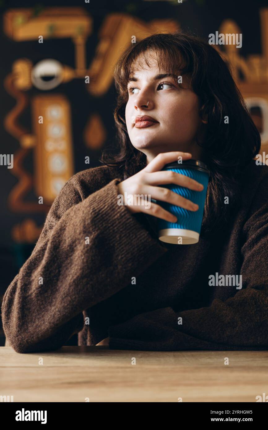 Une jeune femme réfléchie avec un anneau nasal est assise dans un café confortable, tenant une tasse de café bleue. L'arrière-plan présente un décor chaud et flou, créant un rela Banque D'Images