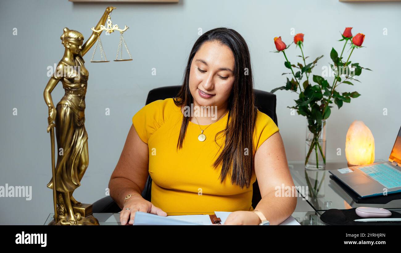 Une avocate latina dans une blouse jaune examine des documents à son bureau, avec une statue d'écailles et de roses, Un ordinateur et une lampe éclairent le bureau moderne s. Banque D'Images