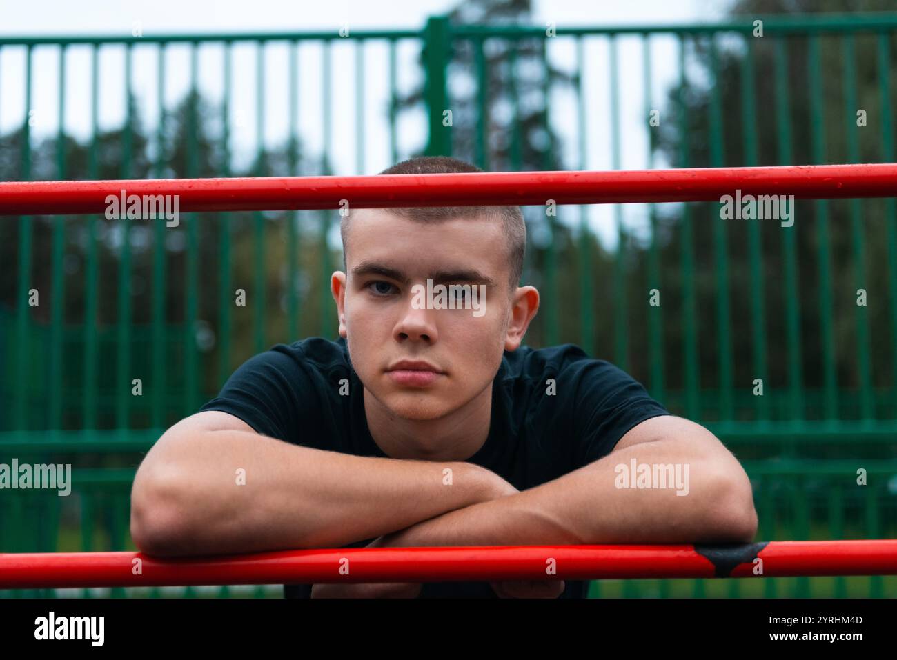 Un jeune homme en chemise noire se penche soigneusement sur une barre rouge dans un parc sportif en plein air. La clôture verte et les arbres créent un arrière-plan serein Banque D'Images