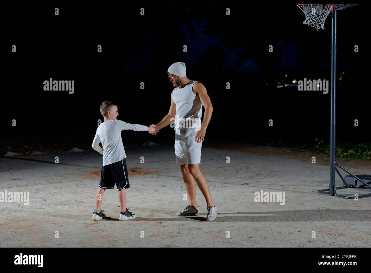 Un père joue au basket-ball avec son jeune fils la nuit dans un parc éclairé par une lumière vive Banque D'Images